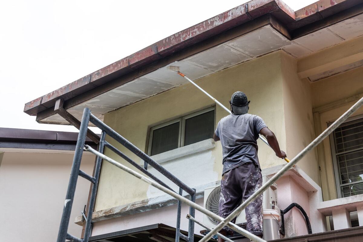 A painter on scaffolding rolling primer onto a stucco wall under the eaves